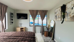 Ein modernes Schlafzimmer mit einem großen Bett, einem Flachbildfernseher, einem Nachttisch aus Holz, einem Spiegel und einem Fenster mit Vorhängen.
