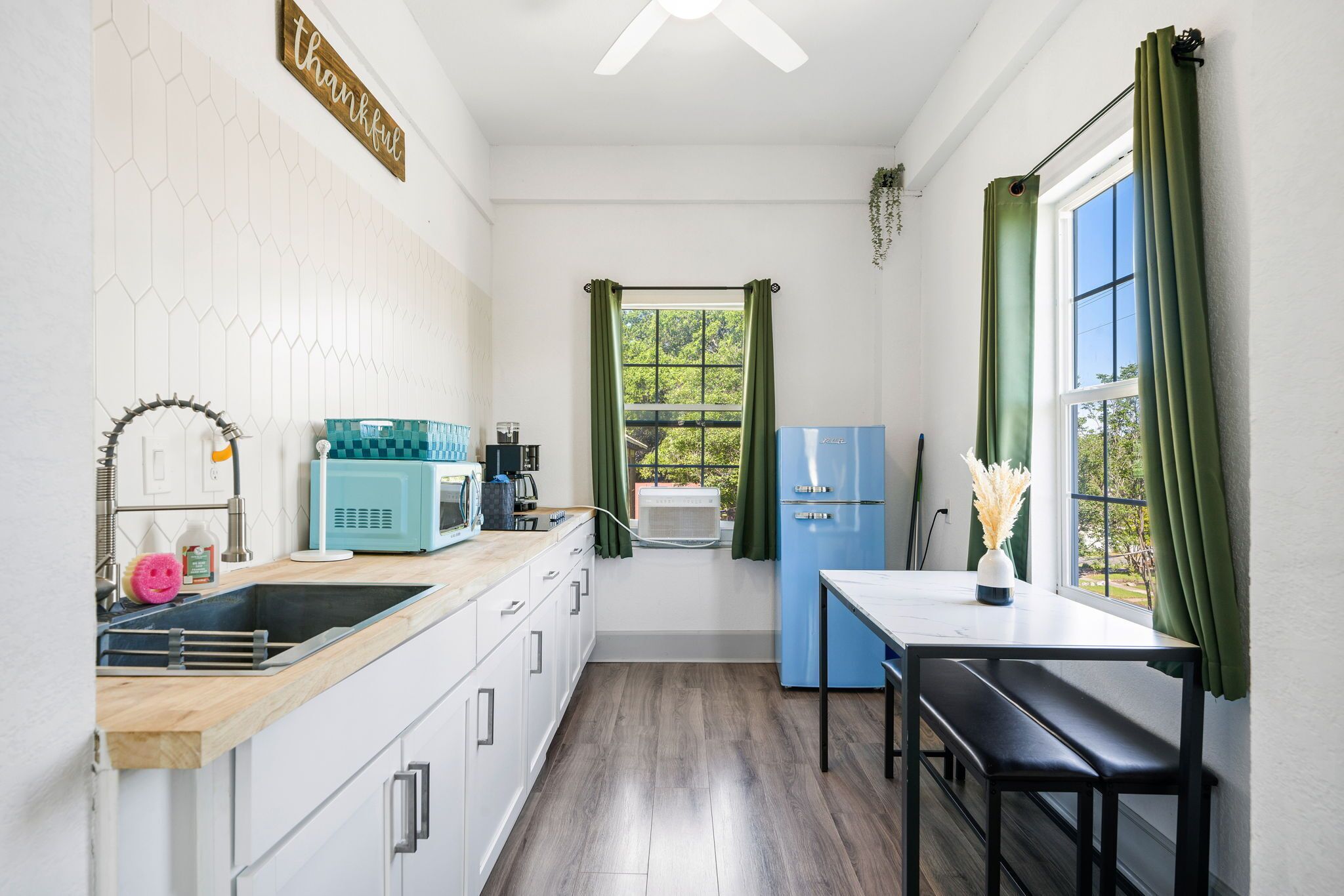 A compact kitchen with a wooden countertop, a blue refrigerator, a sink, and a window with green curtains.