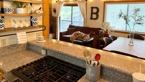 A kitchen with granite countertops, a gas stove, a coffee maker, and a mug on the counter. There is a dining area with a table and chairs, and a living room with a brown sofa and a blanket. - Pet Friendly Condo w/ Hot Tub in Shaver Lake (Shaver Lake)