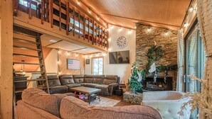 A cozy living room with a sectional sofa, a fireplace, a loft with a wooden staircase, and a stone wall. - Pet Friendly Condo w/ Hot Tub in Shaver Lake (Shaver Lake)