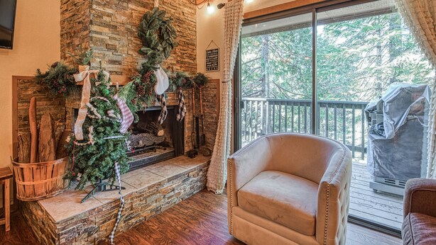 A cozy living room with a stone fireplace, a comfortable armchair, and a large window overlooking a deck. - Pet Friendly Condo w/ Hot Tub in Shaver Lake (Shaver Lake)