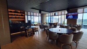 Un restaurant moderne avec des tables et des chaises rondes, un coin bar avec des étagères et de grandes fenêtres donnant sur l’extérieur.