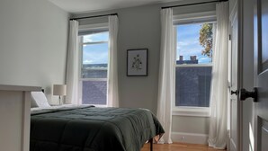A bedroom with a bed, a nightstand, a lamp, and two windows with curtains.