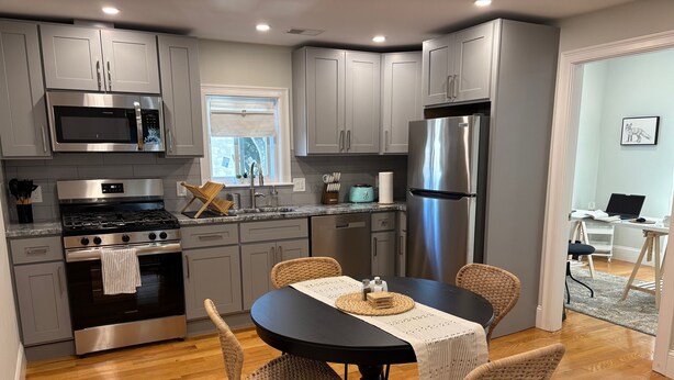 A modern kitchen with grey cabinets, stainless steel appliances, a dining table with woven chairs, and a visible window.