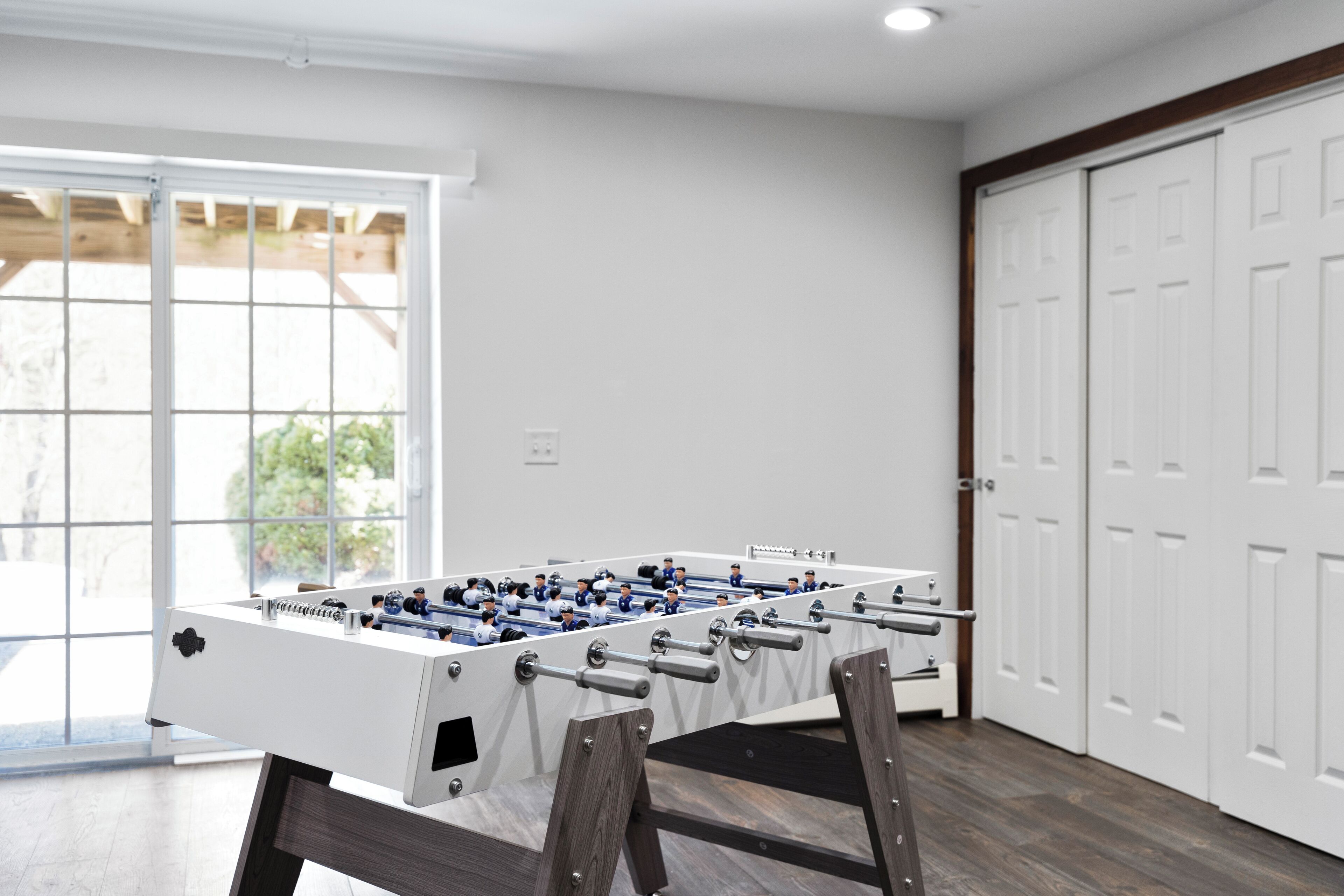 A foosball table in a room with wooden flooring, white walls, and a large window.