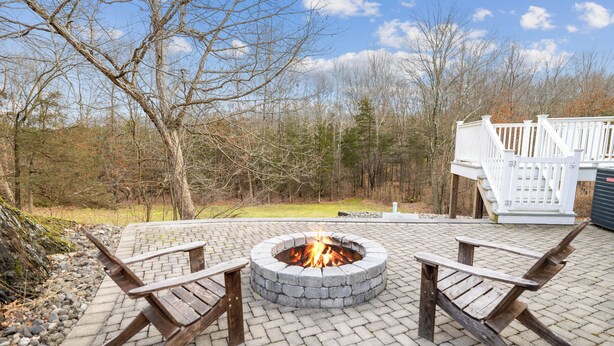 A circular fire pit with two wooden Adirondack chairs, surrounded by a paved patio area with a white deck in the background. - Private 5-Bed Country Estate on 22 Acres (Chester)