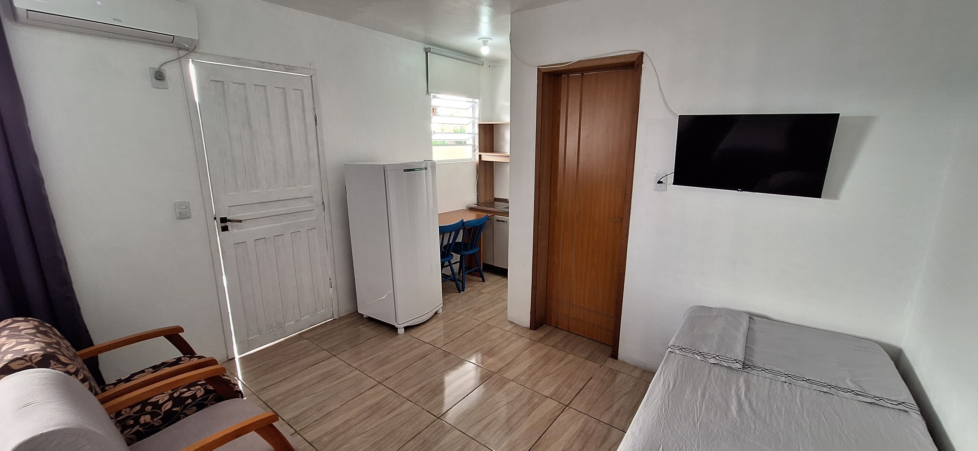 Quarto compacto com cama, escrivaninha, cadeira, geladeira e televisão fixada na parede.