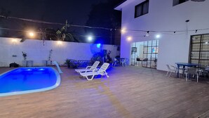 An outdoor swimming pool area with lounge chairs, a table, and string lights. - Pousada Felix (São Gabriel)