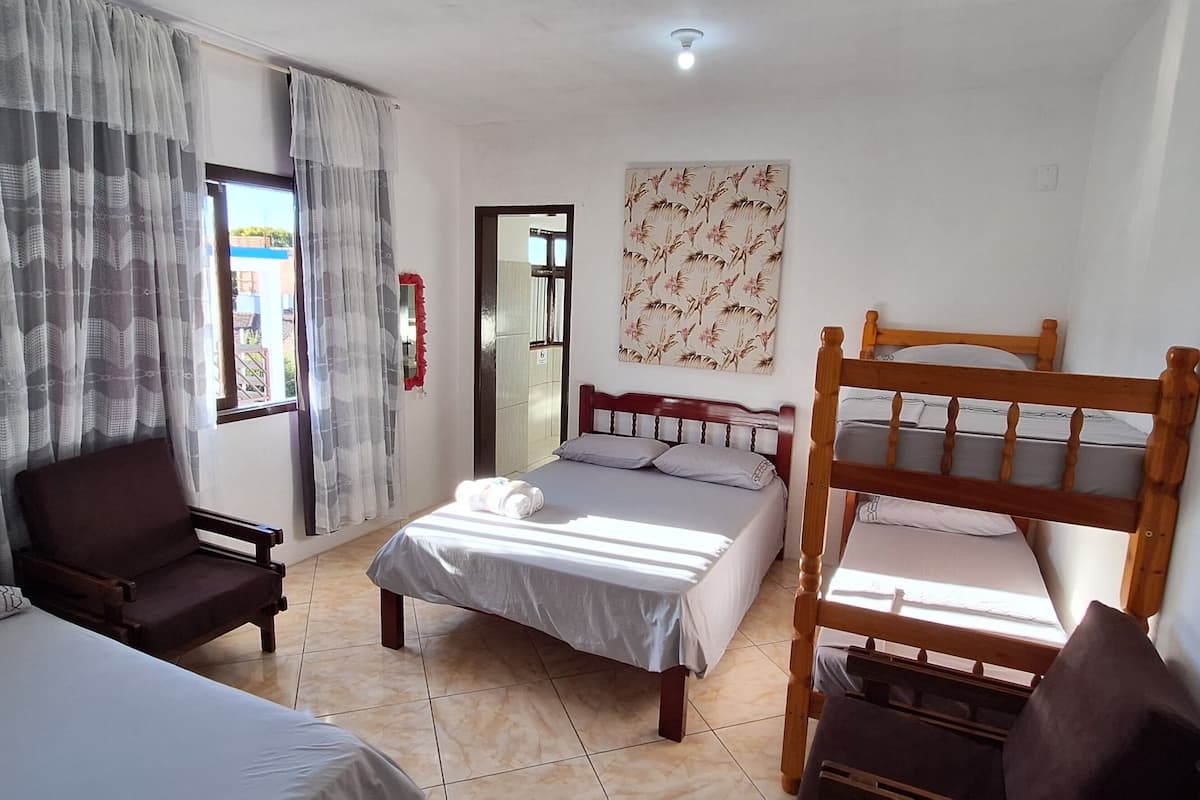 A room with two beds, a wooden bunk bed, a chair, a window with curtains, and a wall with a floral pattern.