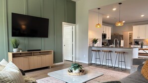 A modern living room with a TV, sofa, coffee table, and a kitchen area with bar stools. - Spacious 4BR Family Retreat- Comfort & Fun for All Ages (Shenandoah)