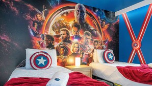 Hotelkamer met twee bedden, elk voorzien van een Captain America sierkussen, een rode deken en een wit kussen. Een nachtkastje met een lamp staat tussen de bedden. Op de muur hangt een groot filmposter.