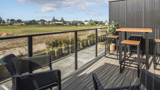 A deck with outdoor seating, a view of a field, and a building in the background. - Waihi Beach Oasis (#B1) (Waihi Beach)