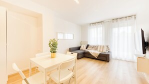 A modern living room with a white sofa, a dining table with chairs, a potted plant, and a large window with sheer curtains.