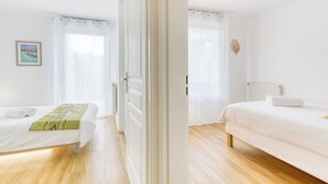A clean, white room with a bed, a bedside table, and a window with sheer curtains.