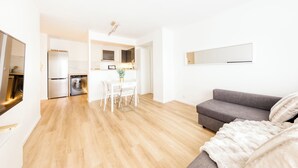 A modern apartment with a grey sofa, a dining area with a table and chairs, a kitchen with appliances, and a large mirror on the wall.