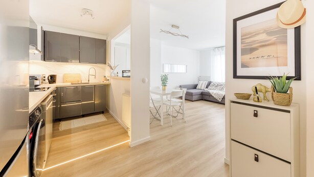 A modern kitchen with stainless steel appliances, a dining area with a white table and chairs, and a living area with a gray sofa.