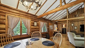 L’intérieur d’une cabane en rondins, avec une table à manger en bois, des chaises, un canapé, et un plafond orné de poutres en bois et d’un lustre.