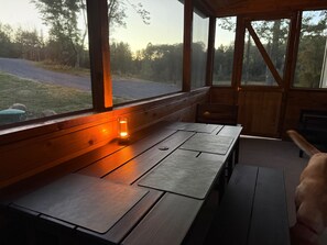 A wooden table with a bench, a small table lamp, and a view of a road and trees through a window. - Craftsman Cottage: Winter Wonderland Cottage — Cozy Pellet Stove & Snowy Lake Views (Iron Mountain)