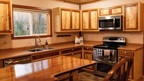 A kitchen with wooden cabinets, a stainless steel microwave, an oven, a sink, and a window with a view of trees. - Craftsman Cottage: Your Perfect Summer Cottage — Swim, Paddle & Relax (Iron Mountain)