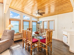 A dining area with a glass table, wooden chairs with red cushions, a ceiling fan, and large windows overlooking a body of water.