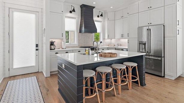 Una cocina moderna con armarios blancos, una isla grande con taburetes de bar, un refrigerador de acero inoxidable y una campana extractora negra.