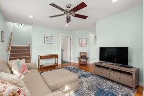 A living room with a sofa, a television on a wooden stand, a ceiling fan, and a staircase leading to another room. - The Ranch: Updated Oceanside home in Duck, Close to Beach w/Views! (Duck)