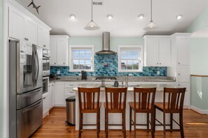 A modern kitchen with white cabinets, a stainless steel refrigerator, a large island with bar stools, and a blue-tiled backsplash. - The Ranch: Updated Oceanside home in Duck, Close to Beach w/Views! (Duck)