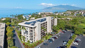 A multi-story hotel with a parking lot, surrounded by greenery and a view of the ocean.