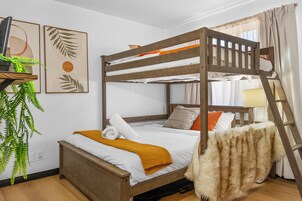 Una cama litera con estructura de madera, ropa de cama blanca y una manta amarilla. Hay una planta en un estante a la izquierda y una escalera a la derecha.