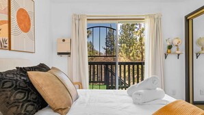 A hotel room with a bed, pillows, a window with curtains, a view of trees, and a balcony with a railing.