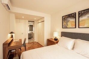 Une chambre d’hôtel avec un lit, des tables de chevet, un bureau et une chaise. Il y a deux tableaux au mur et un appareil de climatisation au plafond.