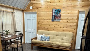 A cozy cabin-style room with a wooden ceiling, a sofa, a dining table with chairs, and a refrigerator.