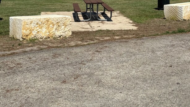 A picnic table with benches, a trash can, and a stone wall in a park by a lake.