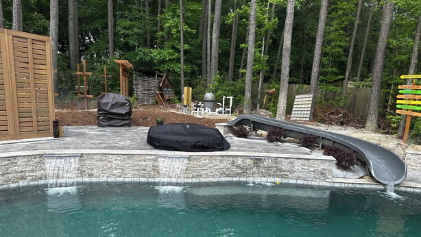 A pool with a waterfall feature, surrounded by a wooden deck and a forested area.