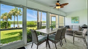 A spacious room with a large window overlooking a lake and palm trees, furnished with wicker chairs and a wooden table.
