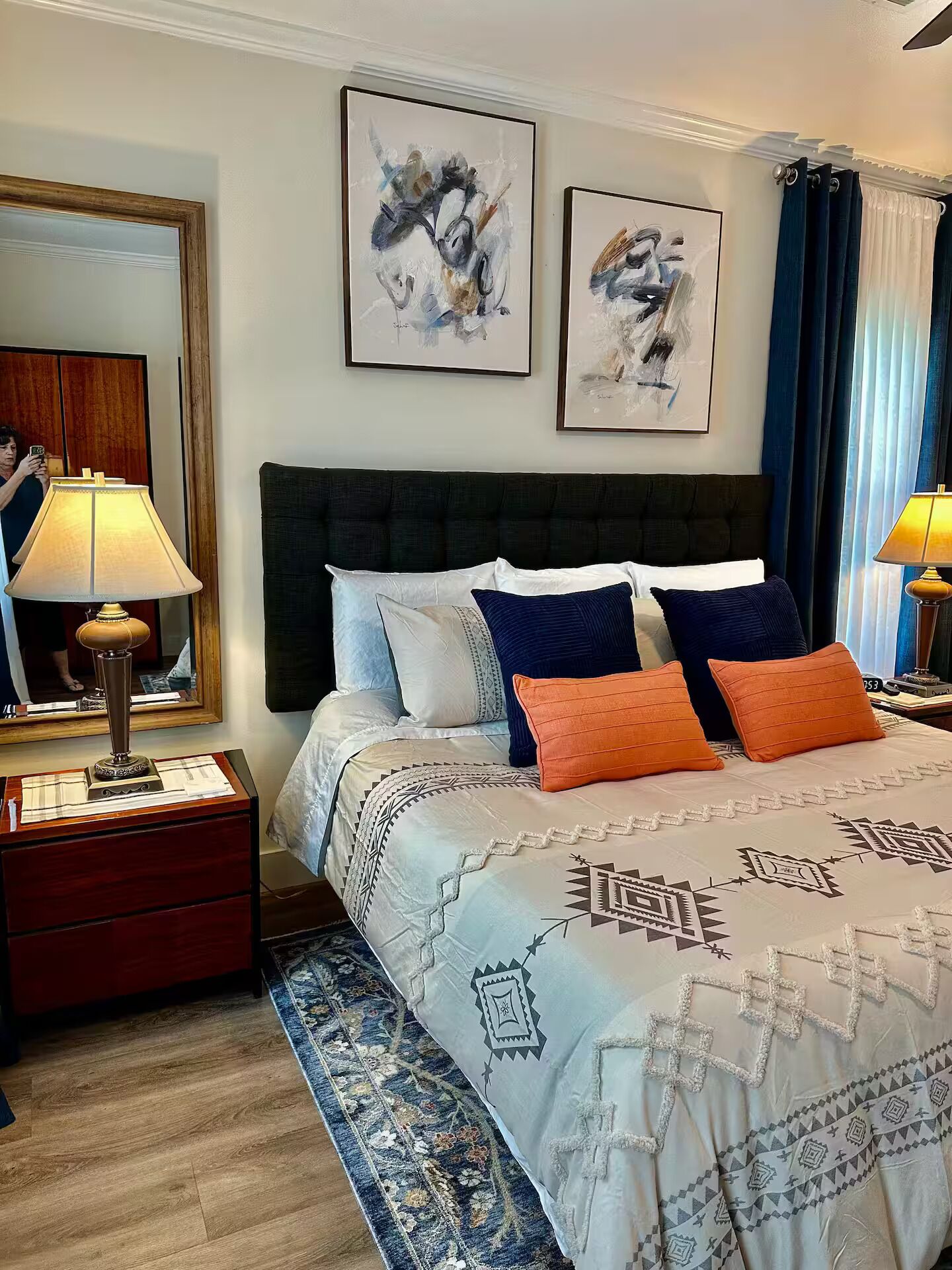 A hotel room with a bed, a nightstand with a lamp, a mirror, and two framed artworks on the wall.
