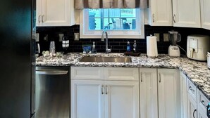 Una cocina con armarios blancos, un refrigerador de acero inoxidable, encimera de granito, lámpara de araña y ventana con vistas al exterior.