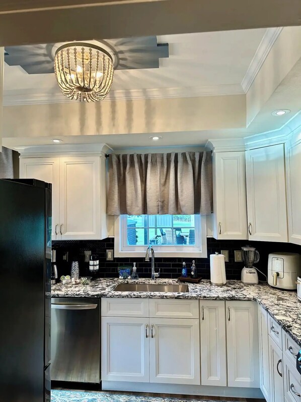 A kitchen with white cabinets, a stainless steel refrigerator, a granite countertop, a chandelier, and a window with a view of the outdoors. - The Blue Heron - Lake View near Music Park (Live Oak)