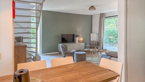 A modern living room with a wooden dining table, white chairs, a grey sofa, a TV, and a balcony with a view of greenery. - Beautiful home in Hoeven with WiFi (Hoeven)