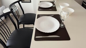 Mesa de jantar posta com pratos brancos, talheres e copos, acompanhada por cadeiras pretas.
