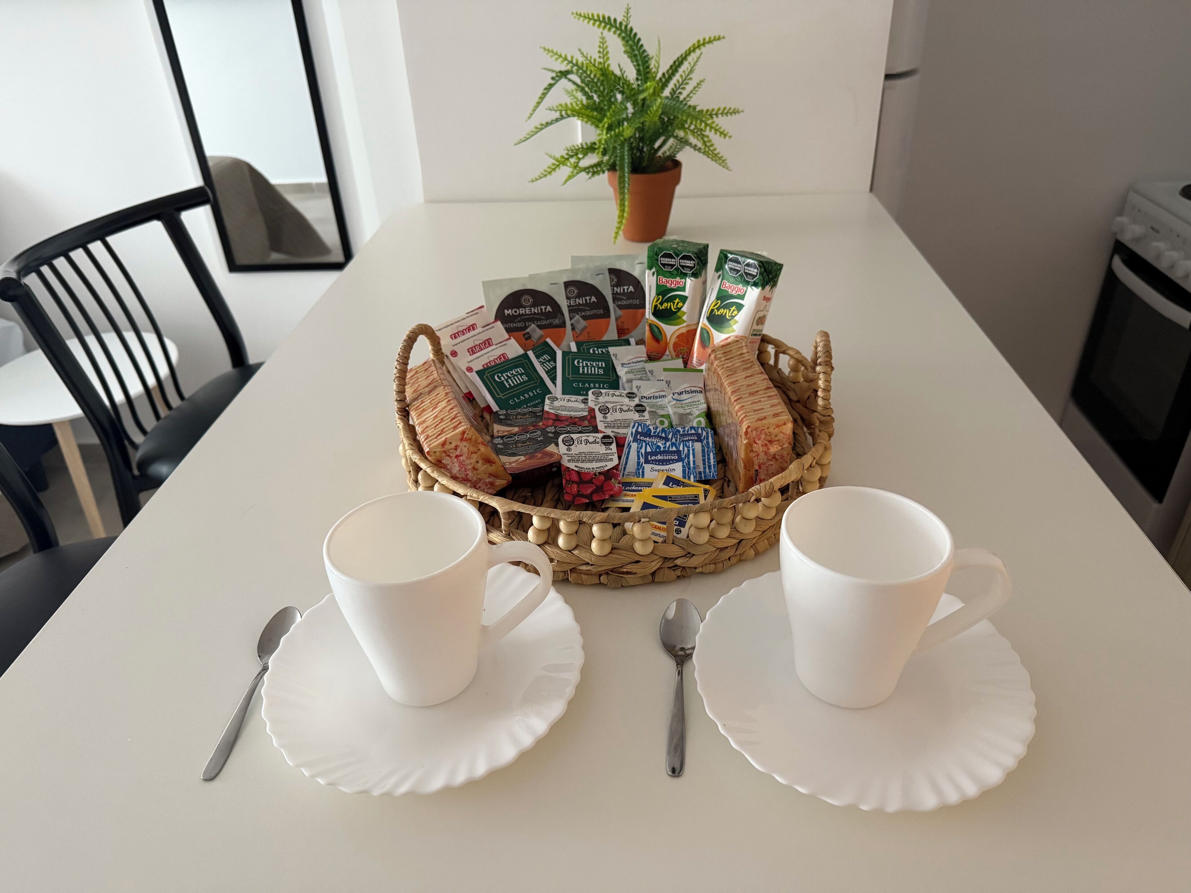 A table with a basket of food items, two white cups, and a potted plant.