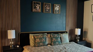 A modern bedroom with a dark blue wall, a wooden slatted ceiling, and a bed with patterned bedding and multiple pillows. There are three framed pictures on the wall and two bedside lamps.