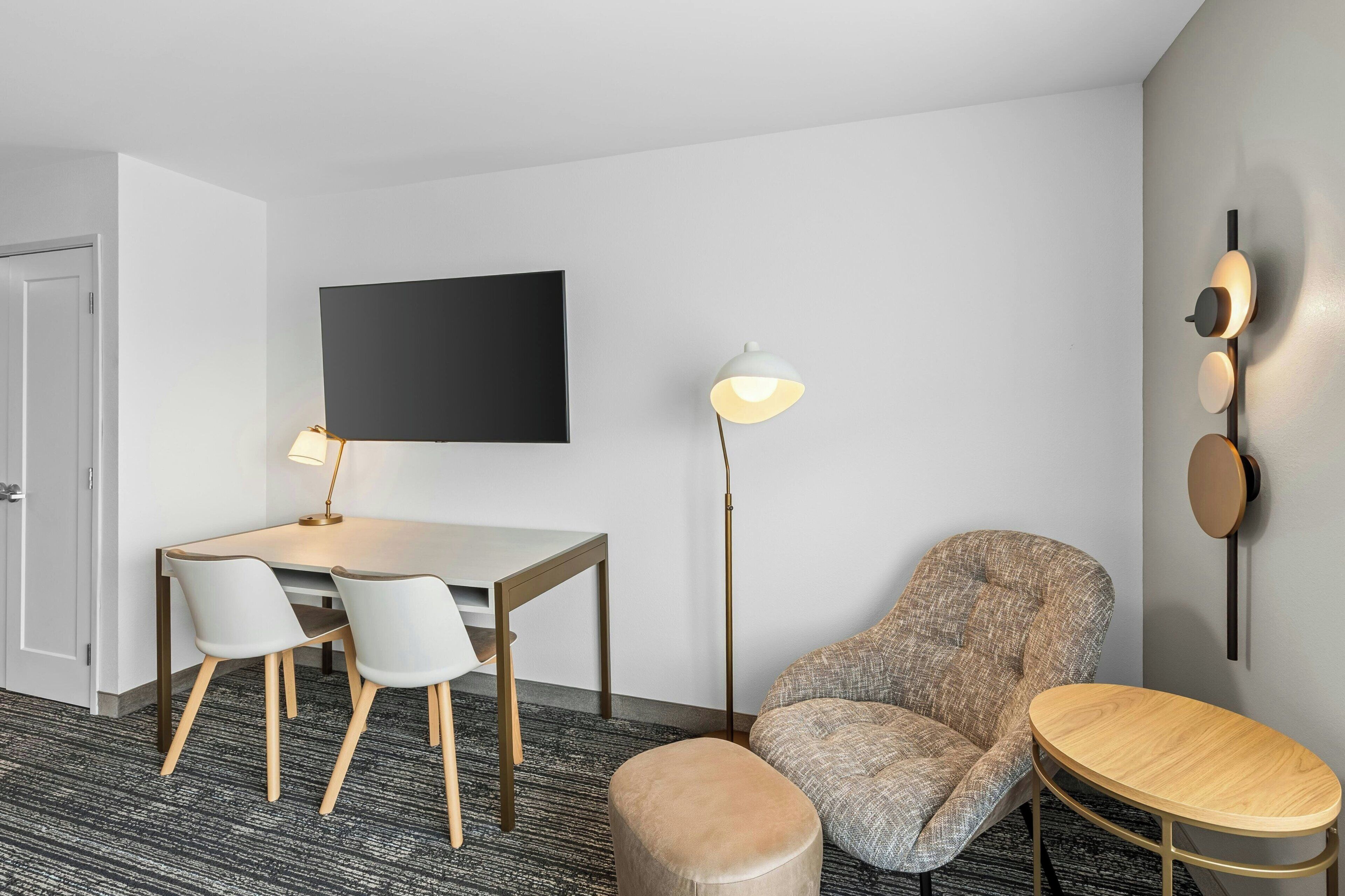 A modern hotel room with a flat-screen TV, a dining table with chairs, a round coffee table, a patterned armchair, a floor lamp, and a wall-mounted light fixture.