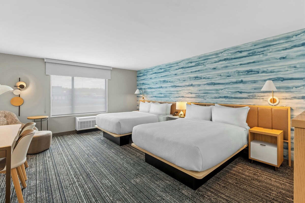 Habitación de hotel con dos camas, un escritorio, una silla y un ventanal grande.
