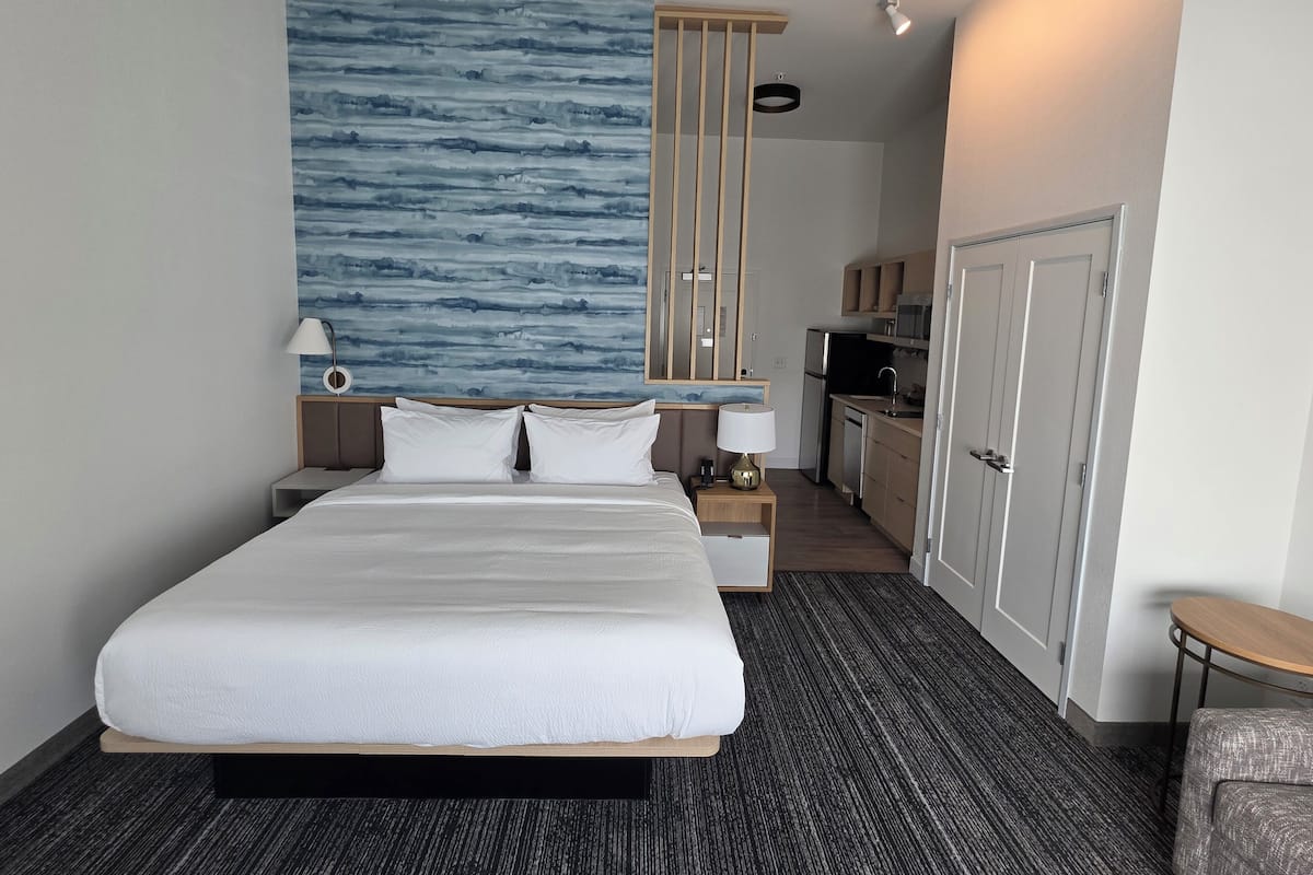 A hotel room with a large bed, a nightstand, a desk, a kitchenette, and a bathroom.