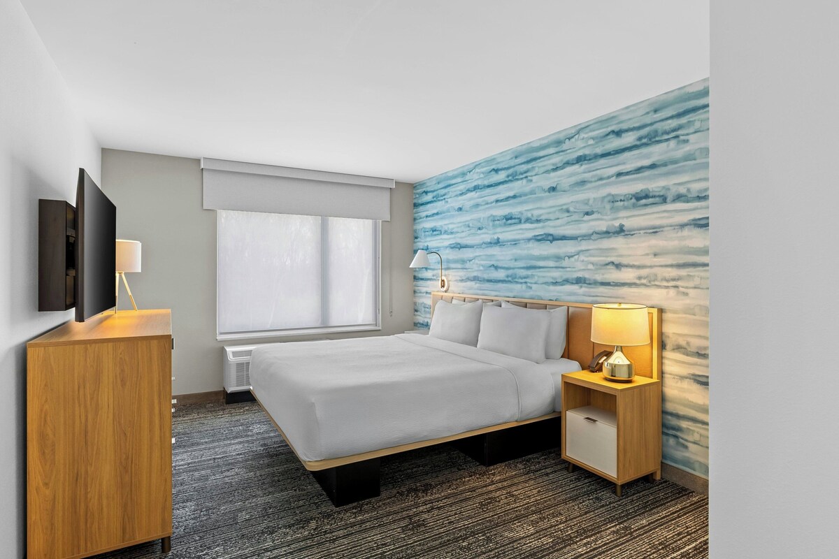 Habitación de hotel con cama, mesita de noche, lámpara, televisor y pared con un diseño abstracto azul.