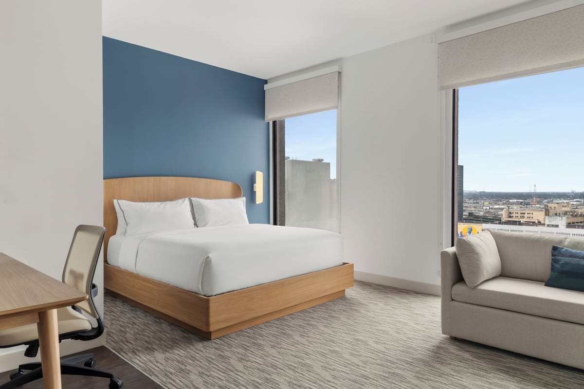 A hotel room with a bed, a desk, a chair, a sofa, and a large window with a city view.