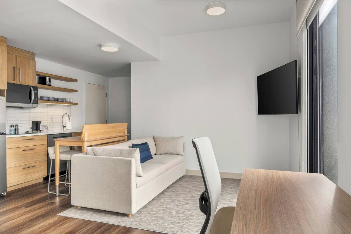 A modern, compact living space with a kitchenette, a sofa, a dining table, and a TV mounted on the wall.