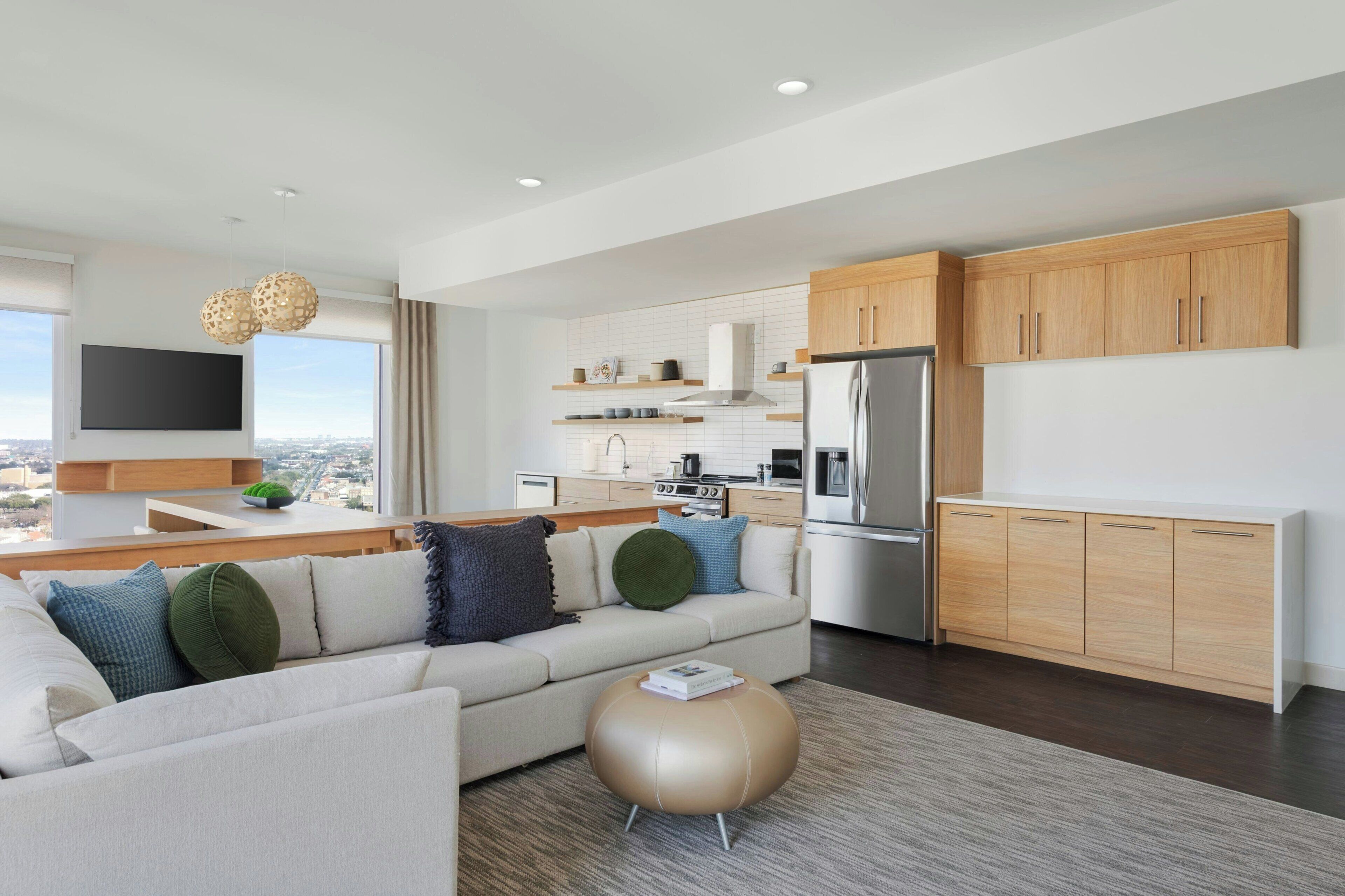 A modern living room with a sofa, a round ottoman, and a kitchen with wooden cabinets and a stainless steel refrigerator.