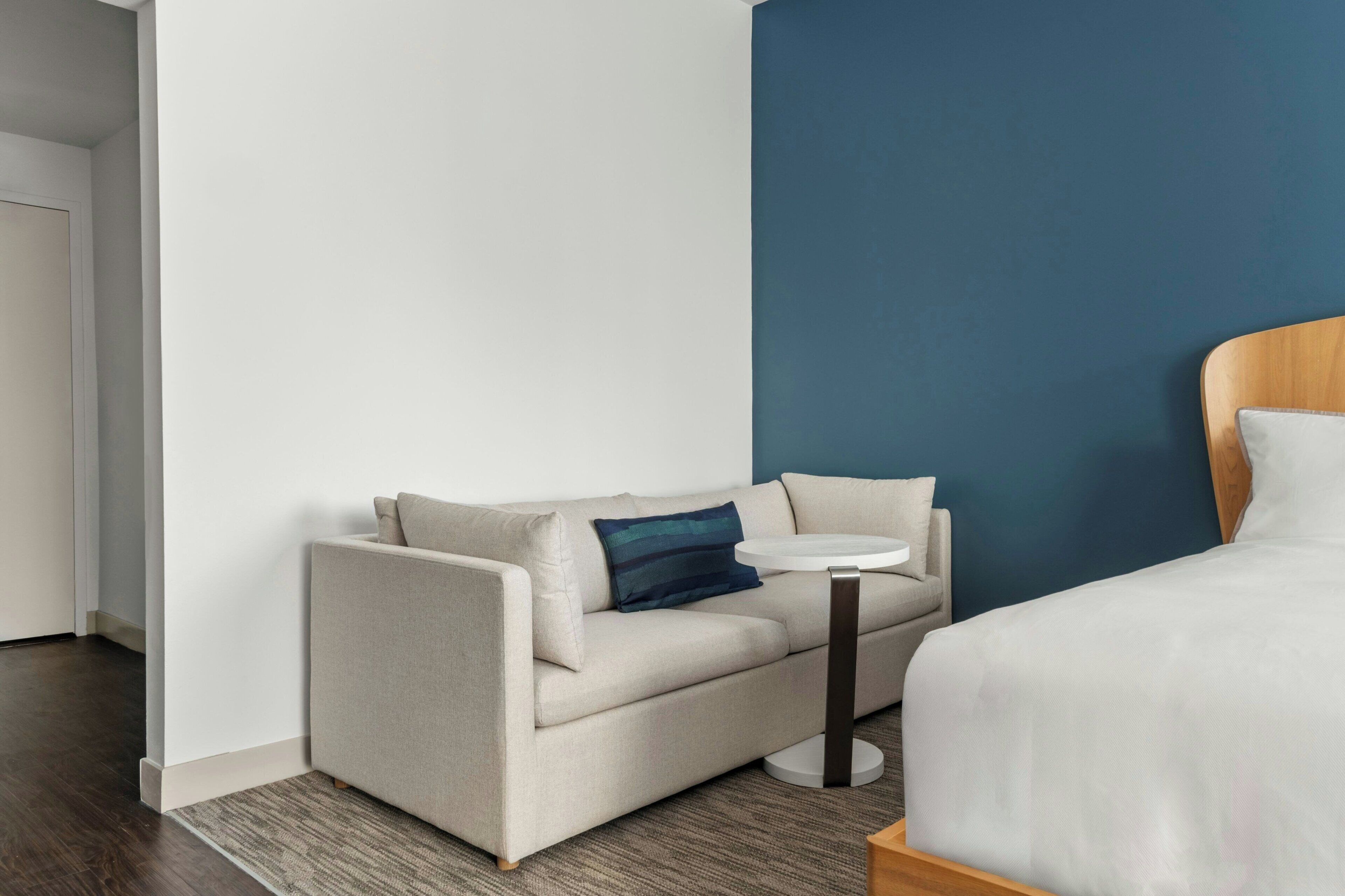 A hotel room with a beige sofa, a small round table, a bed with white bedding, and a blue accent wall.
